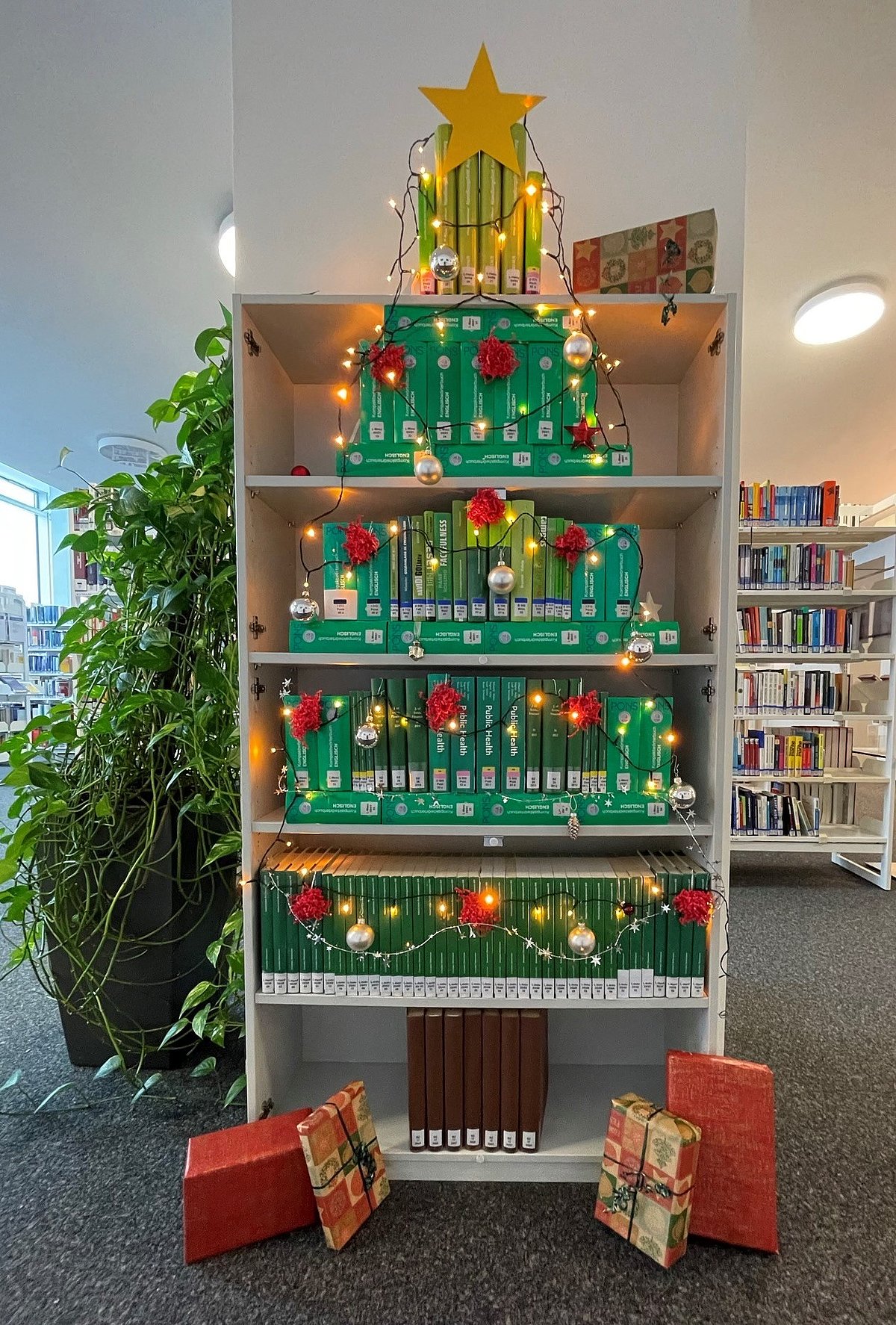 Ein Bücherturm in Form eines Tannenbaumes aus grünen Büchern mit Lichterkette und Kugeln dekoriert.